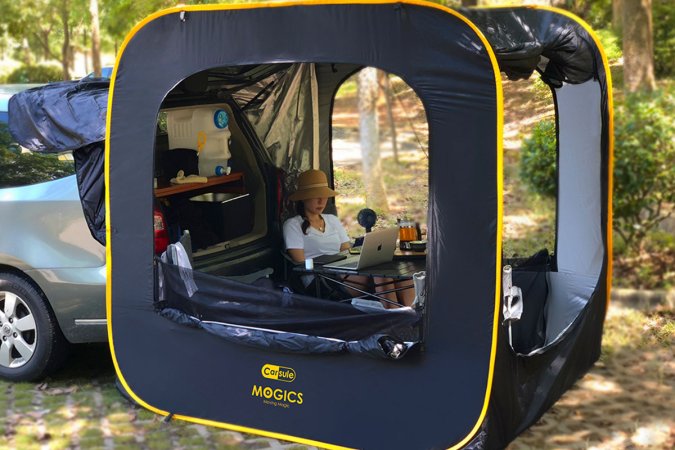 A person working on their laptop while sitting in a cube-shaped tent behind their car in the woods