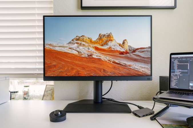  A BenQ SW272U monitor sits on a white desk with the Hotkey Puck and laptop stand next to it.