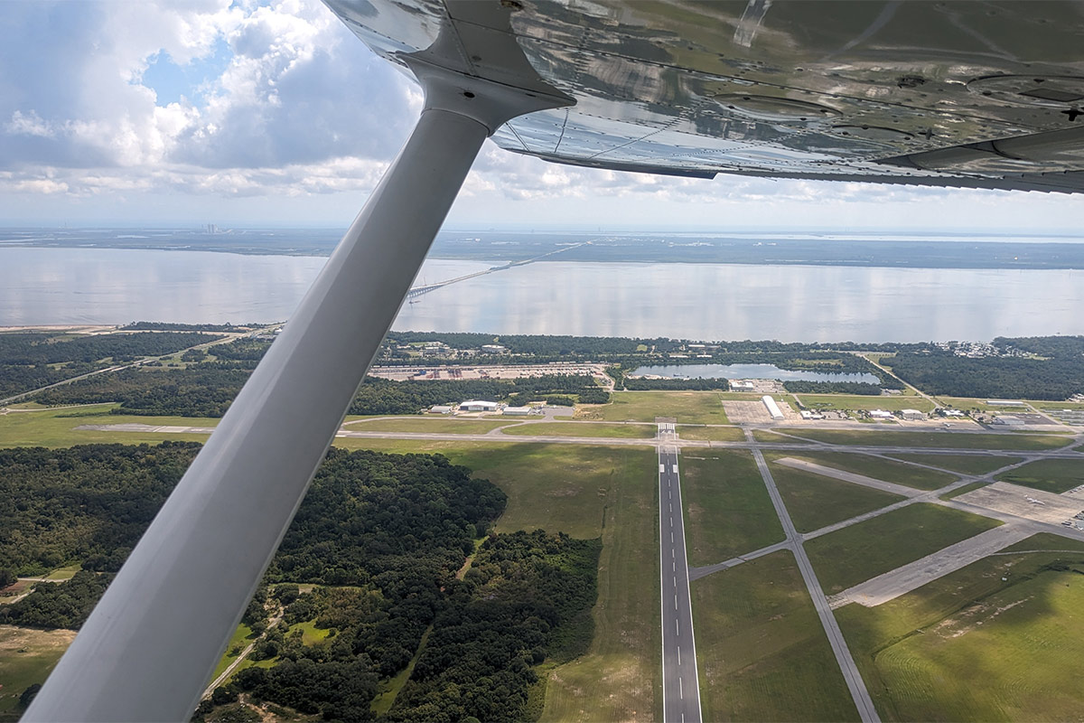 A photo taken from the window of a small airplane under the wing with an airfield and ocean in the distance.