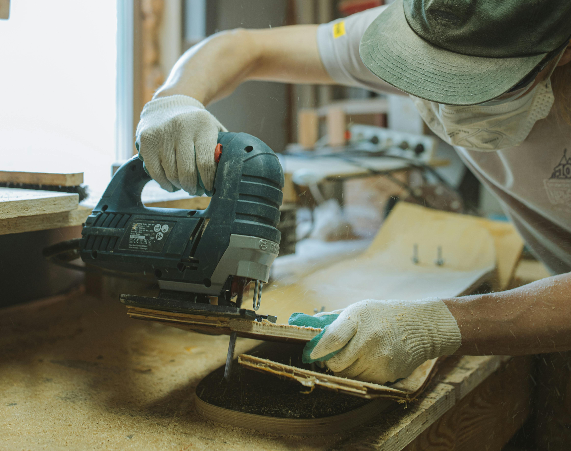 A person wearing a green baseball cap using a jigsaw to cut the edge of a skateboard deck out of a piece of wood.