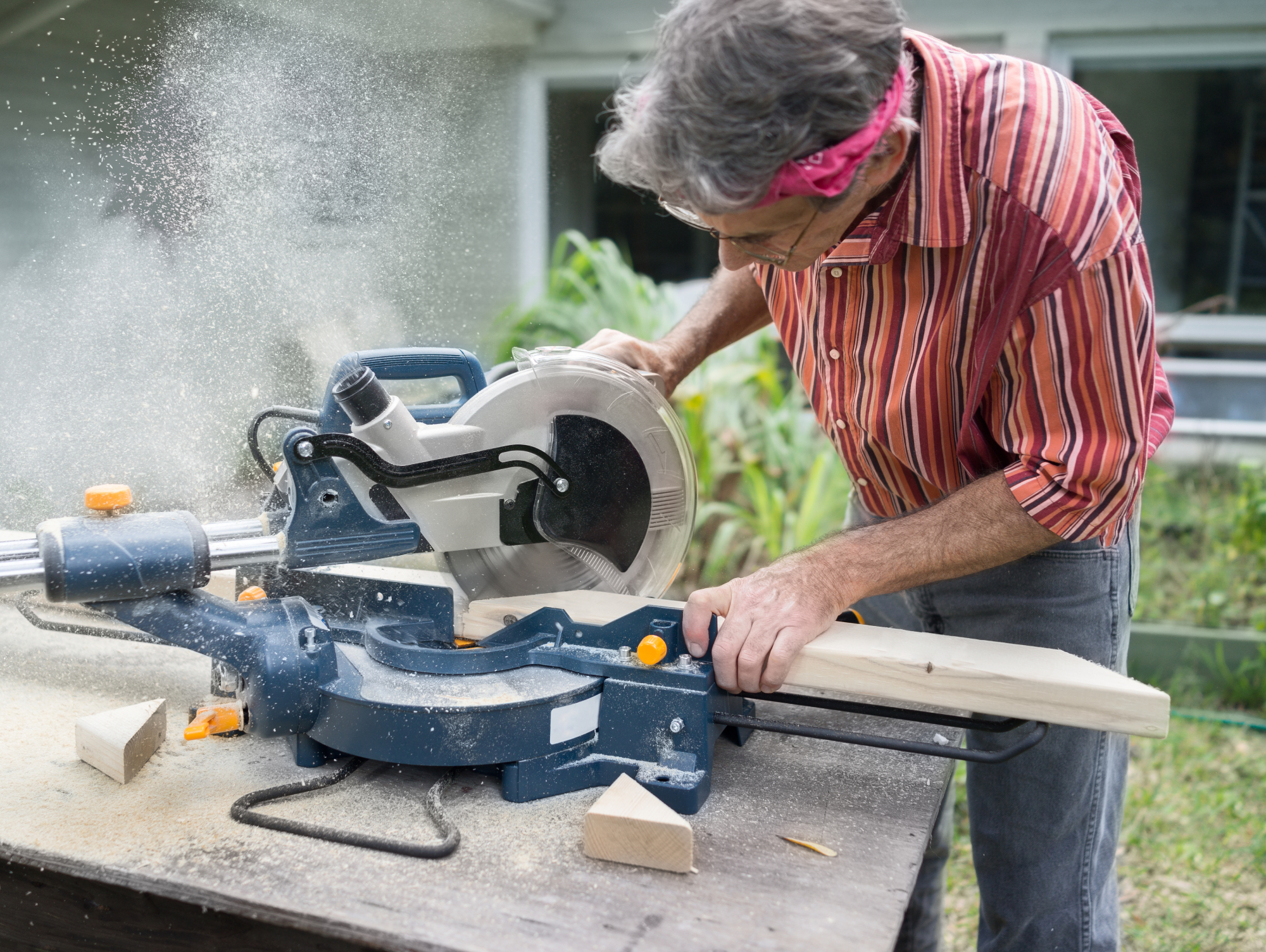 A gray-haired man cutting a piece of wood on an angle while using a miter saw outside.