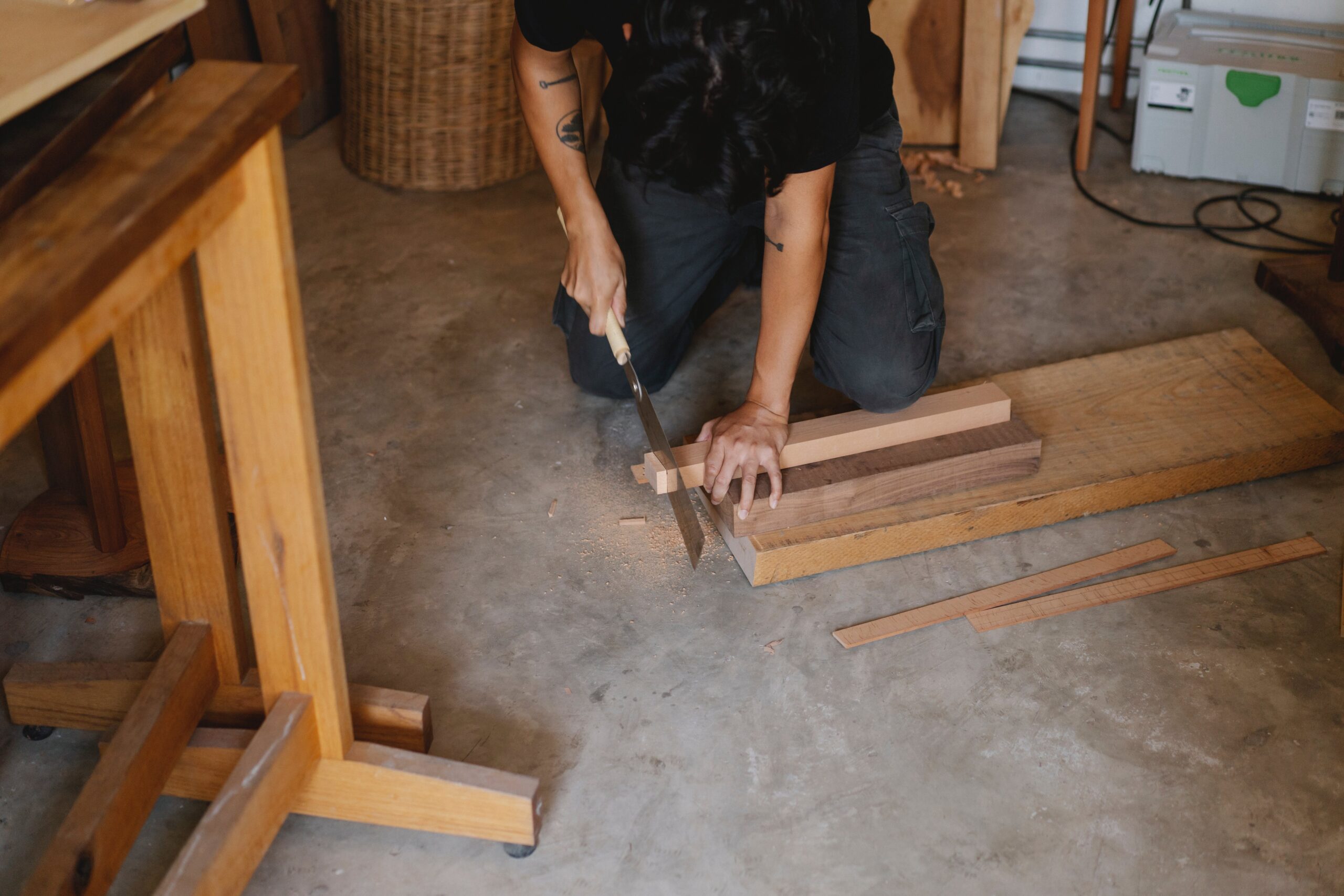 A man kneeling on the floor of a woodshop, using a pull saw to cut through a small piece of wood.