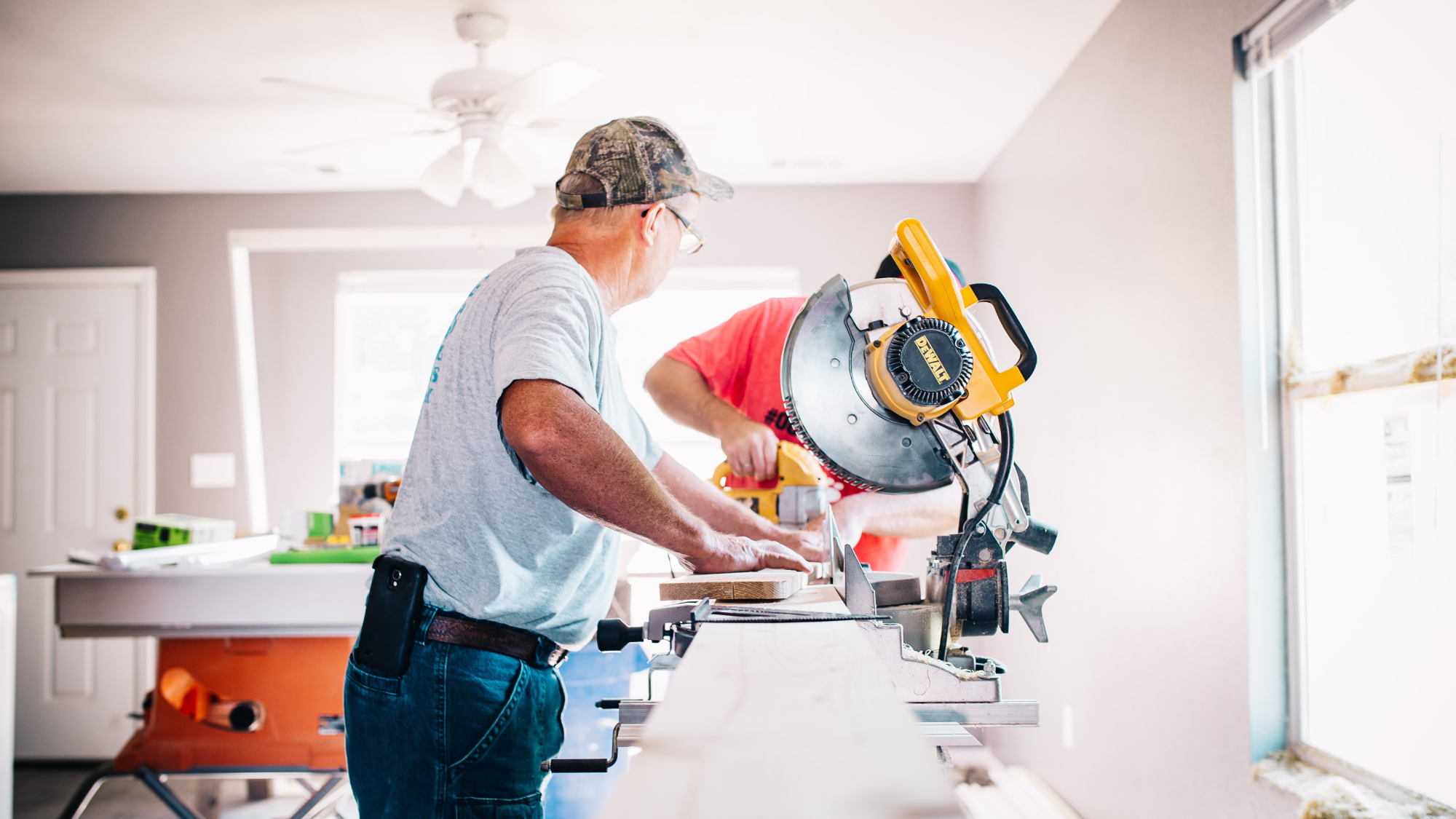 Two older men working in a well-lit white room, using two different types of saws to cut wood: a jigsaw and a miter saw.