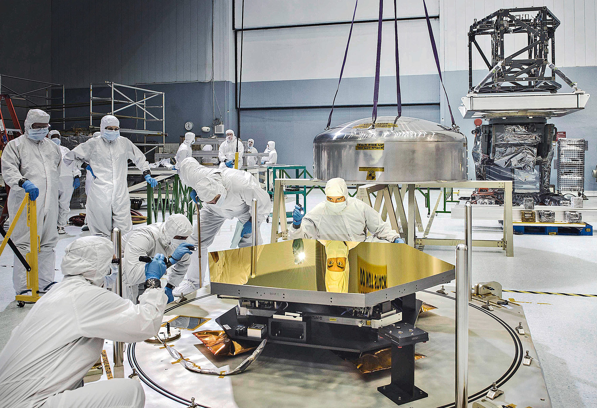 A team of scientists in a large room look at a gold mirror for the James Webb Space Telescope.