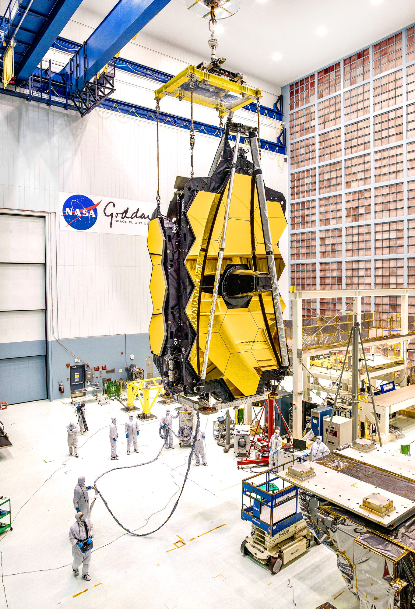 The gold mirrors of the JWST hang in a large room at NASA Goddard with groups of scientists working below.