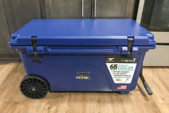  A blue ORCA 65-Quart Insulated Rolling Cooler sits on the floor in front of some kitchen cabinets.