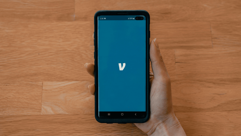 A hand holding a phone over a wooden table. The phone screen is blue and shoes the logo for the Venmo app.