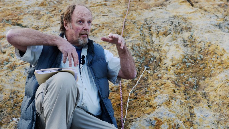 Paleontologist Martin Lockley sits in front of a rock with dinosaur claw marks and shows how dinosaurs must have clawed into the rock during a mating ceremony where they made pseudo nests for potential mates.