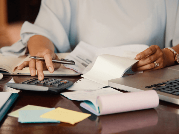 A person sitting at a desk with a calculator, notebook, and laptop set up in a messy fashion.