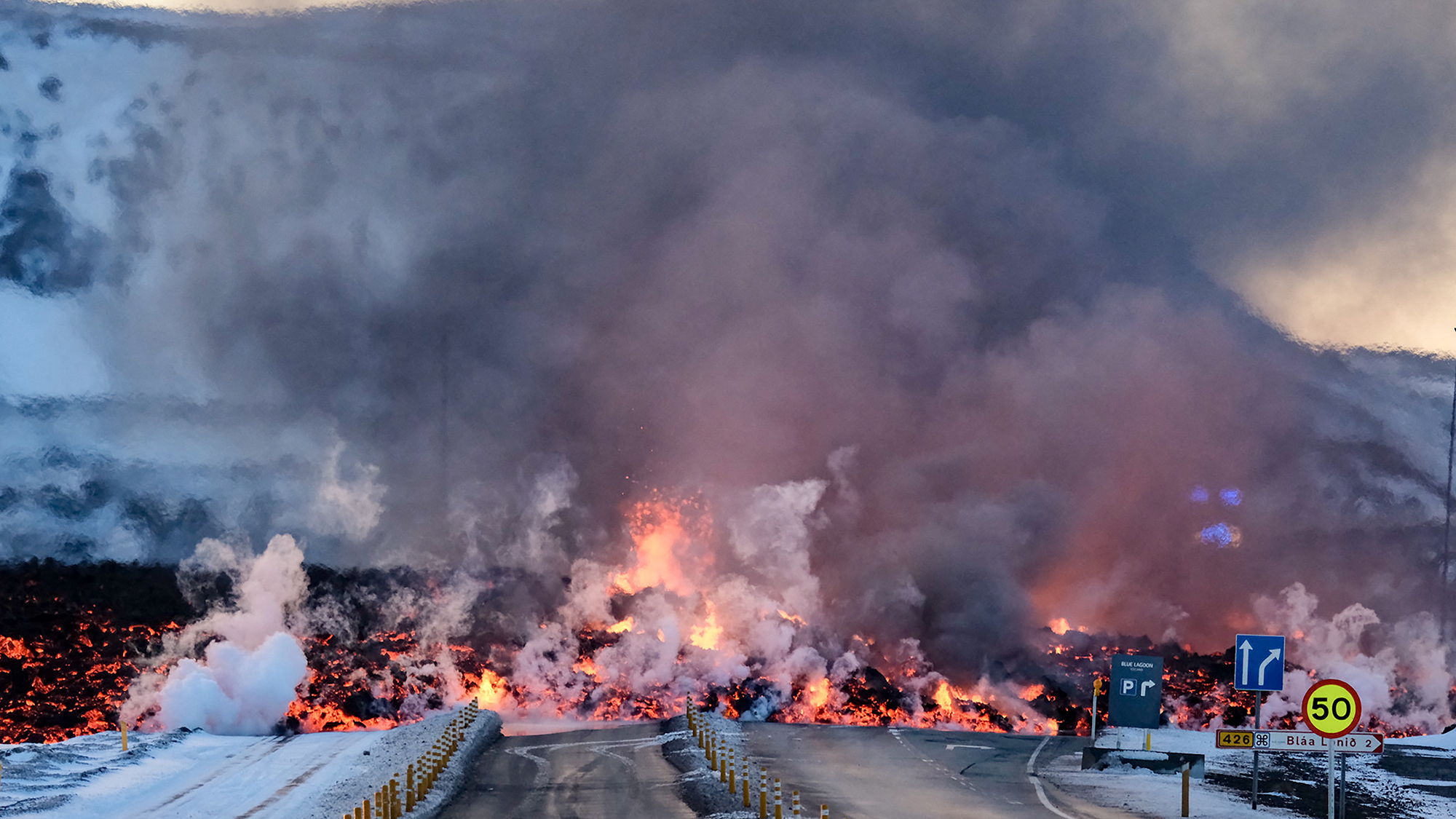 Molten lava overflows the road leading to the Blue Lagoon geothermal spa, a popular tourist destination in western Iceland. Orange lava covers a dark road, as gray smoke rises.