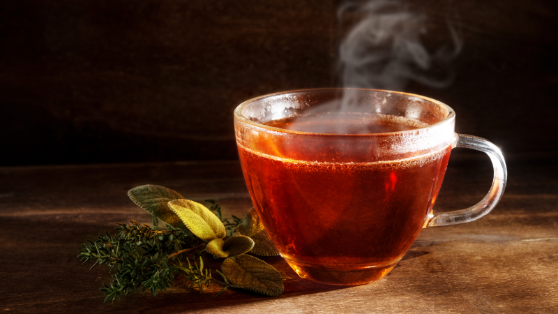 A clear glass tea cup with steaming brown tea sits on a table next to a green tea leaves.