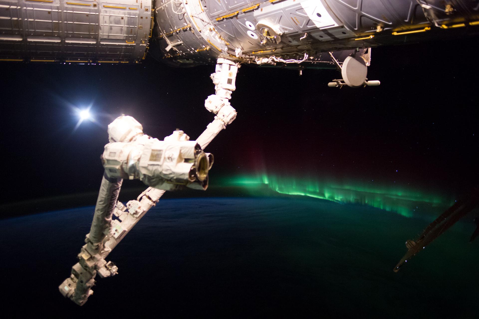 the arm of a space craft in the foreground with a green aurora in the background