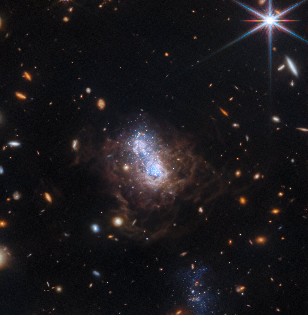 A black background is dotted with numerous white elliptical and red spiral galaxies. A dwarf irregular galaxy dominates the image, with a bright area of ​​white and blue stars at its center that look like two distinct clumps. This area is surrounded by brown dust filaments. A companion galaxy can be seen in the lower center of the image, which looks like a cluster of blue stars. In the upper right corner is a very prominent bright star with eight long diffraction spikes.