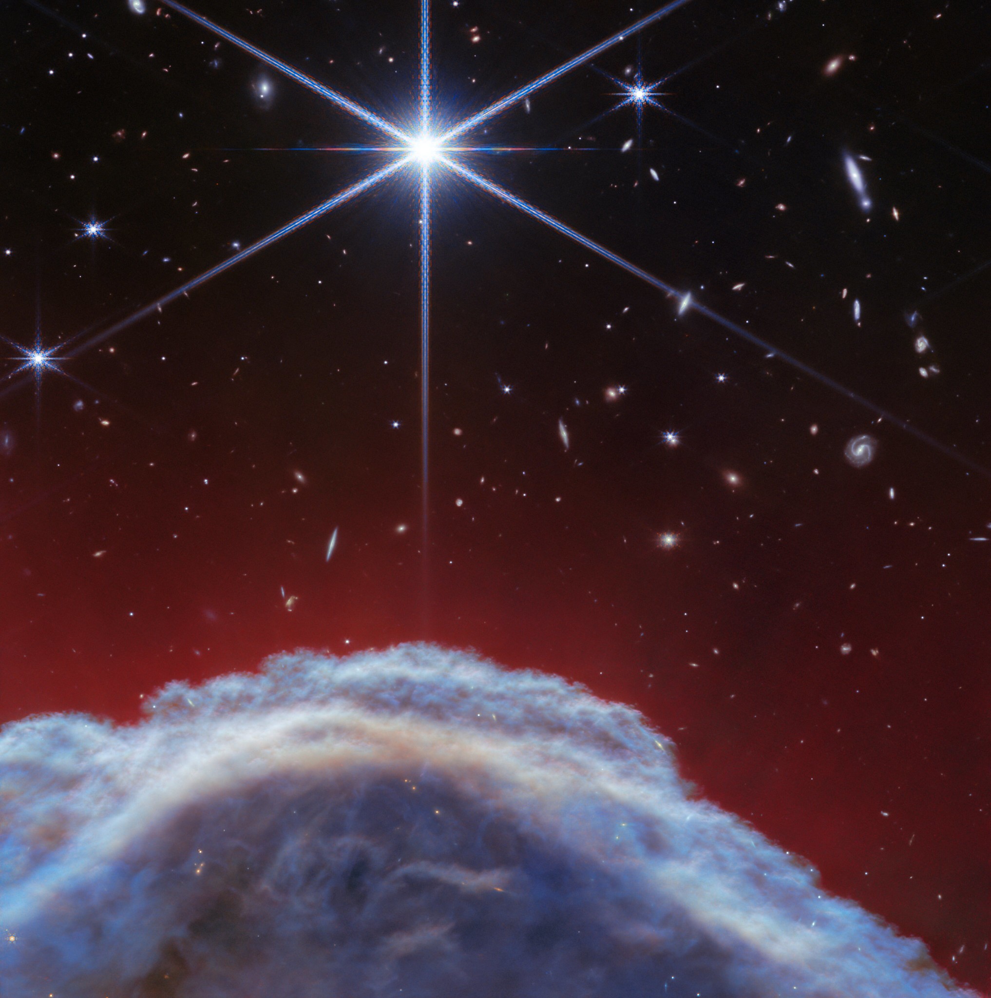 About a third of the way up, a lumpy dome of blue-gray clouds rises.  Above this, striped, translucent red strands brush up about halfway across the image.  The top half of the image is the black background of space with one prominent, bright white star with Webb's 8-point diffraction peaks.  There are other stars and galaxies scattered throughout the image, although very few can be seen through the thick clouds at the bottom and all are considerably smaller than the largest star.