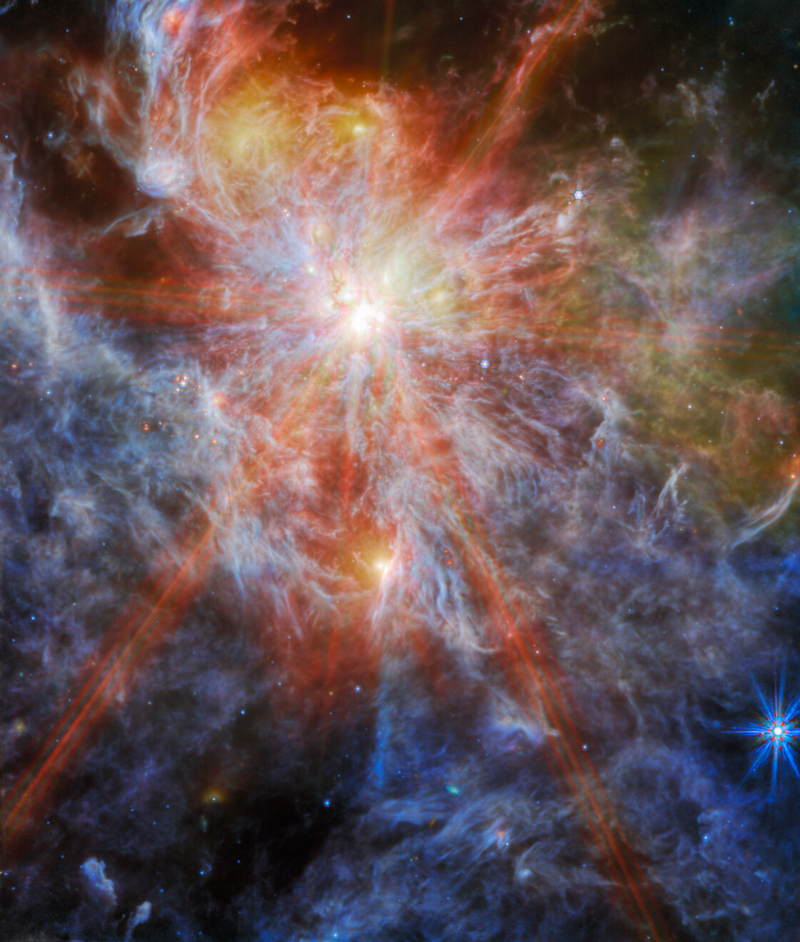 A bright young star, located in the upper left quadrant, shines through layers of wispy white and blue clouds on a dark background.  The star is surrounded by thick orange points in an eight-pointed pattern, spanning most of the image.  A patch of greenish-yellow clouds appears at the top right of the image.  There are a few other bright spots visible as glowing yellow dots among the clouds, as well as another bright star with smaller blue diffraction peaks in the lower right corner.