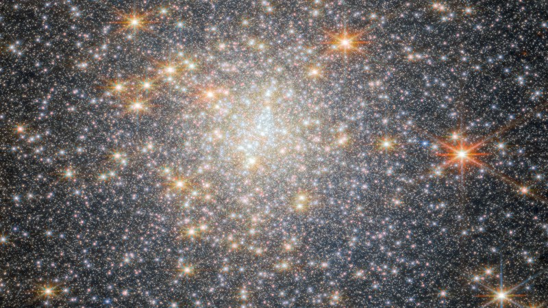 A spherical collection of stars which fills the whole view. The cluster is dominated by a concentrated group of bright white stars at the center, with several large yellow stars scattered throughout the image. Many of the stars have visible diffraction spikes. The background is black