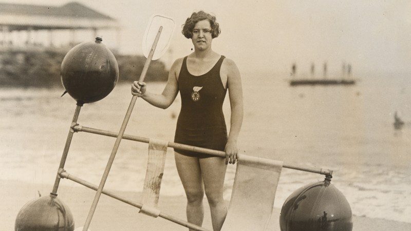 a black and white photograph of swimmer gertrude ederle. she is standing on a beach in a one piece swimsuit holding an oar.