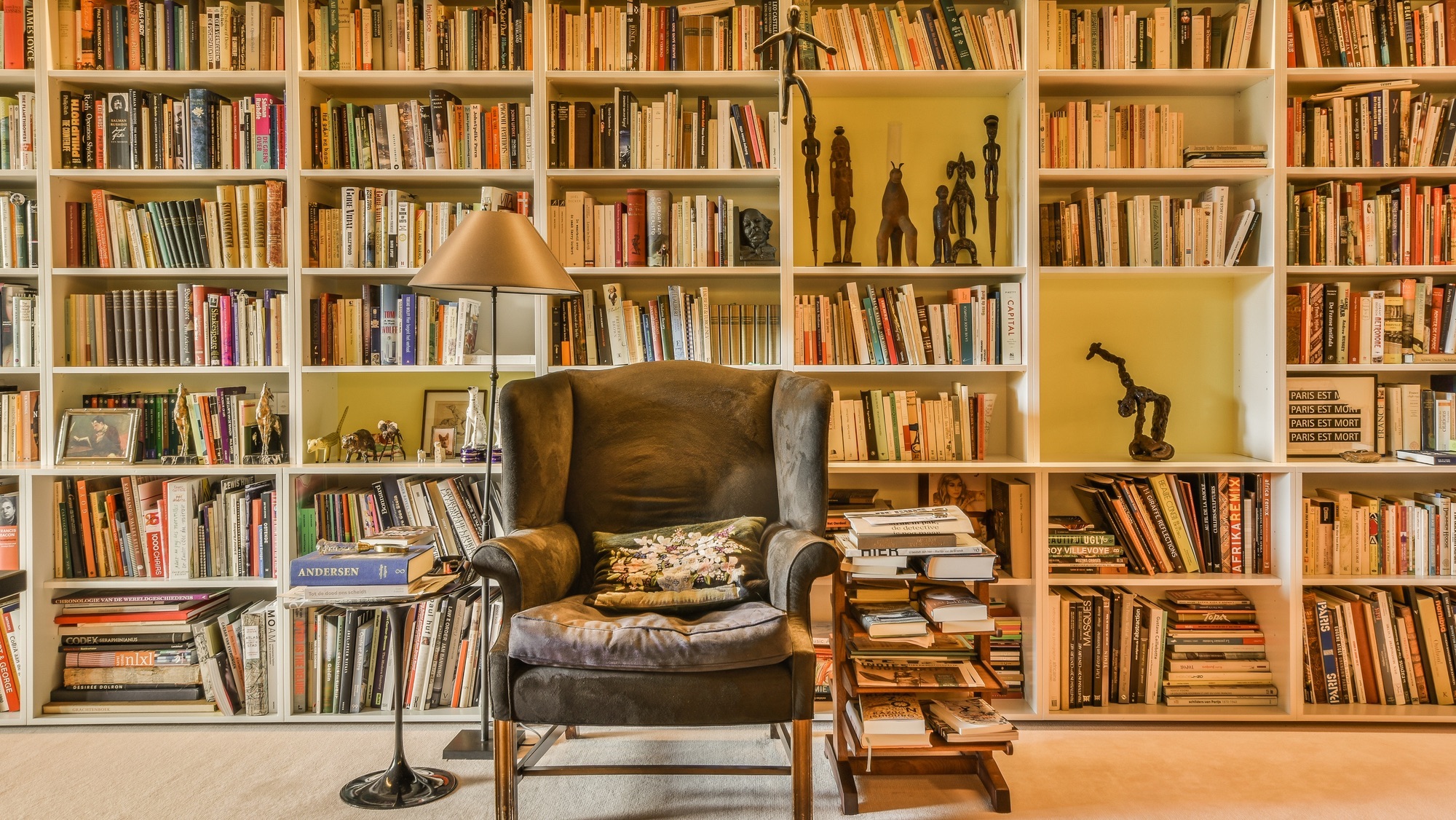 Amsterdam, Netherlands - 10 April, 2021: a chair in front of a book shelf with many books on it and a lamp sitting next to the chair
