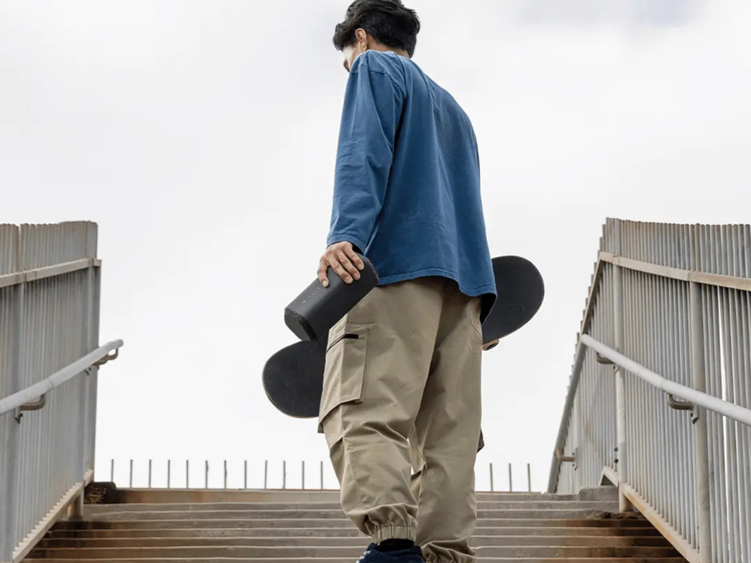 A person carrying a skateboard and a Sony XE300 Portable Bluetooth Speaker.