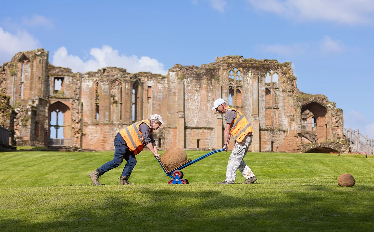 two workers in orange vests carry a large stone on a dolly in front of a medieval castle 