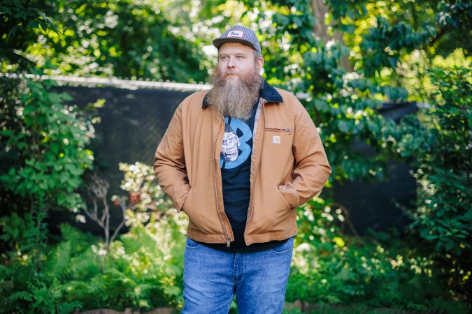  A man standing in a carhartt jacket in front of some trees with a hat on