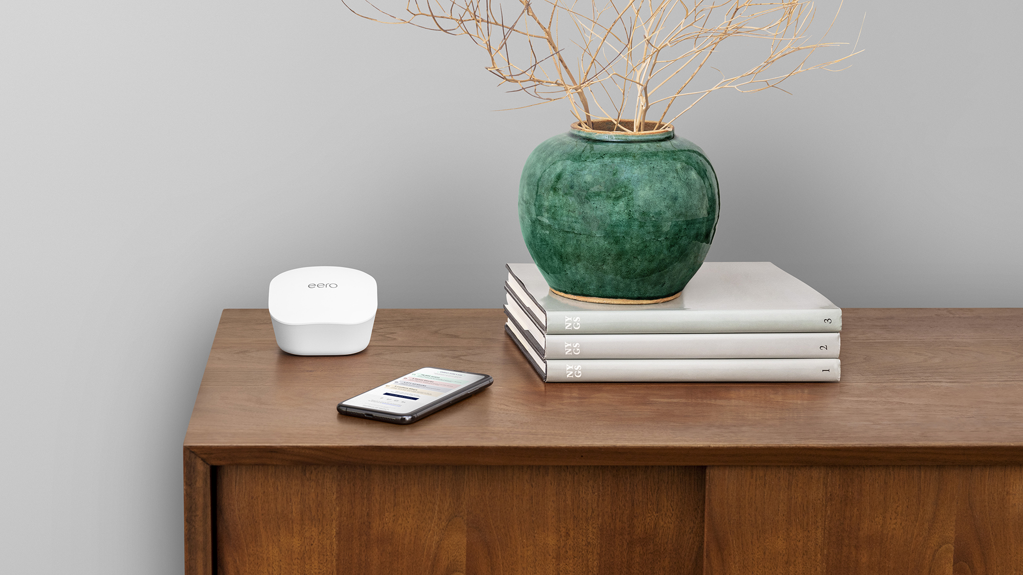 a credenza with a pile of books, vase, and eero device on top