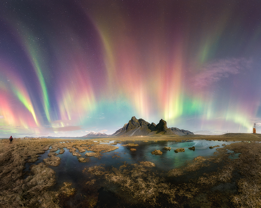 a rainbow aurora over a mountain near a lake.  a person is standing on the left