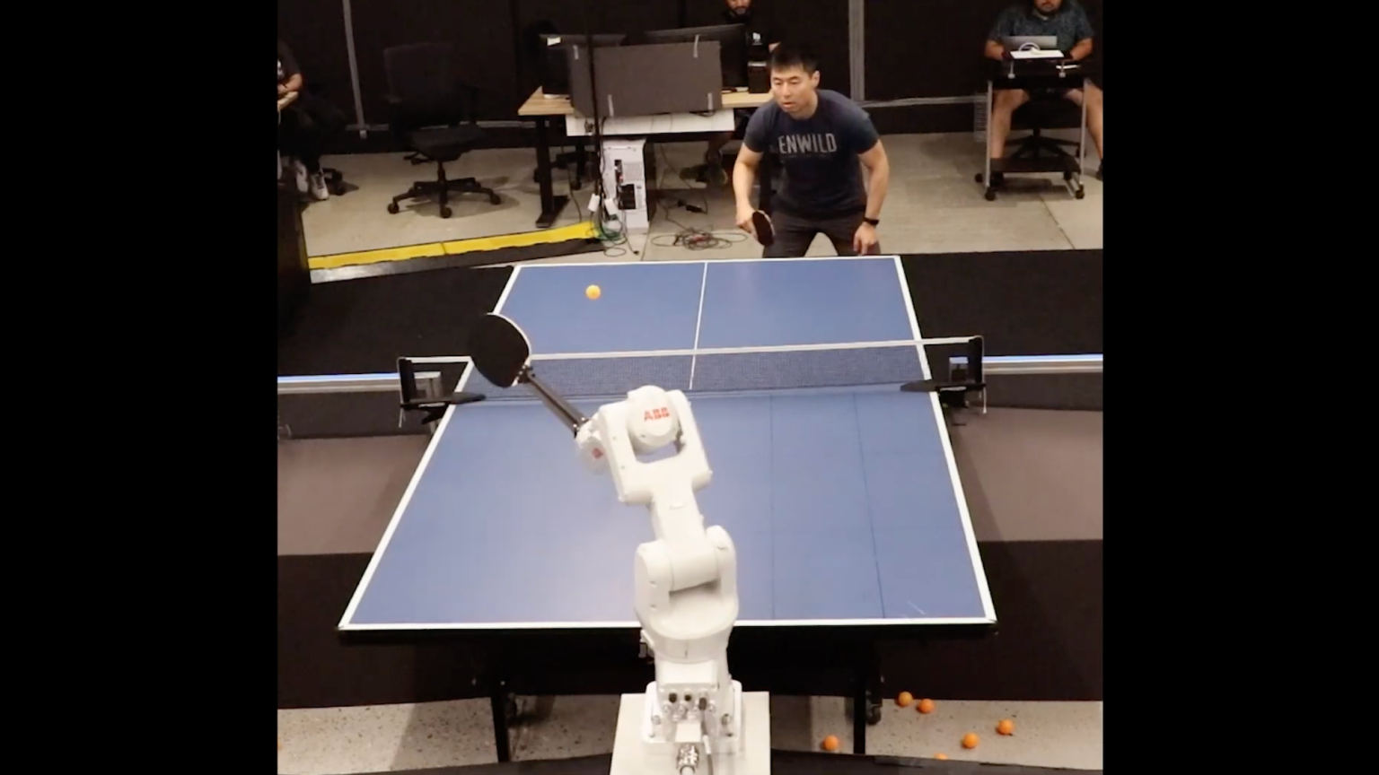 Un brazo robótico equipado con una pala de ping pong se enfrenta a un jugador humano en una partida de tenis de mesa. El brazo robótico, marcado con el logotipo de ABB, está en posición para devolver la pelota mientras el jugador humano, concentrado, se prepara para el siguiente golpe. En el fondo, se pueden ver otras personas sentadas observando o trabajando en computadores, en lo que parece ser un entorno de laboratorio o investigación.