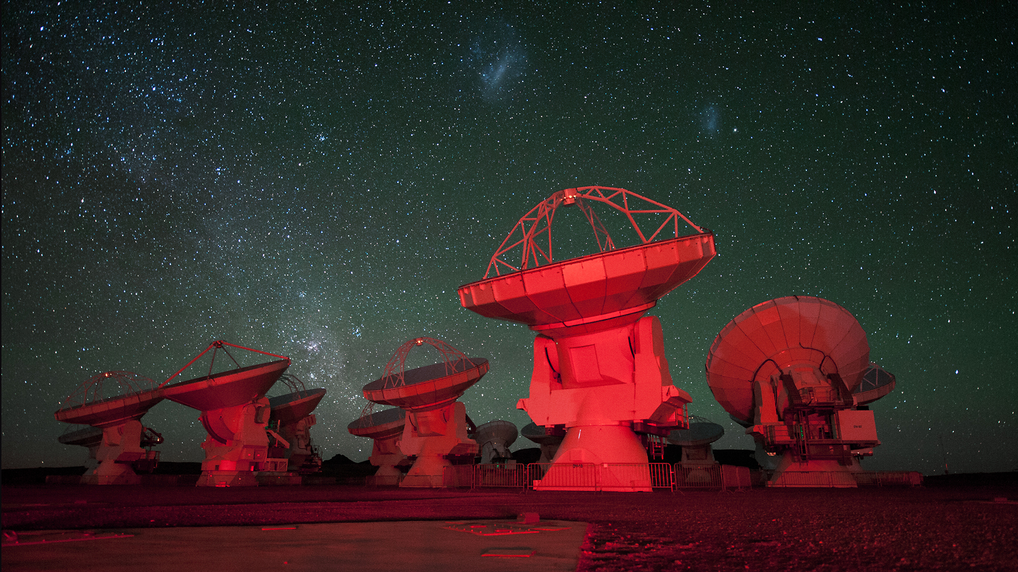 ALMA antennae bathed in red light. in the background there is the southern Milky Way on the left and the Magellanic Clouds at the top.