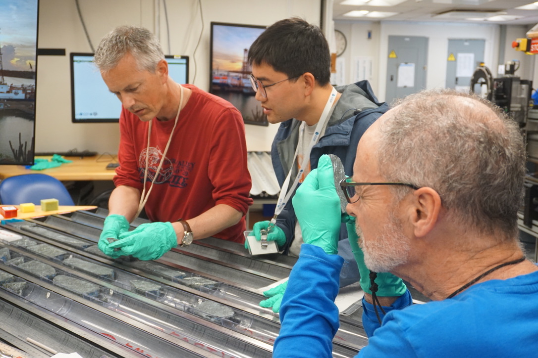 three scientists in a lab analyzing a grey sediment core taken from the earth's mantle