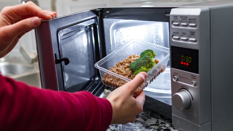 Using the microwave oven to heat food. Woman's hand puts plastic container with broccoli and buckwheat in the microwave