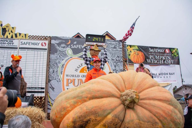 man stands behind large pumpkin with scarecrow