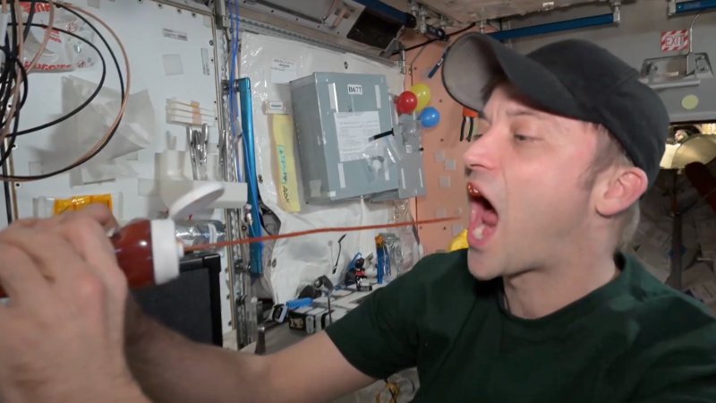 a man wearing a NASA hat aboard the International Space Station squirts ketchup into his mouth. the ketchup comes out in one long stream