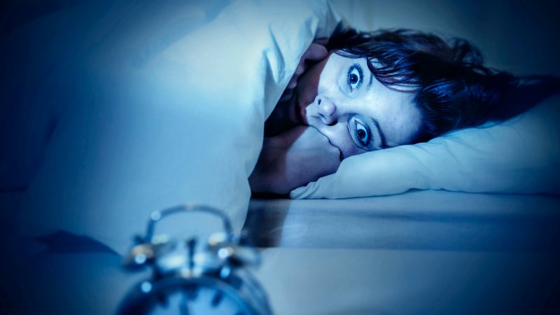 young woman in bed with alarm clock and eyes opened suffering insomnia and sleep disorder thinking about his problem on dark studio lighting in sleeping and nightmare issues