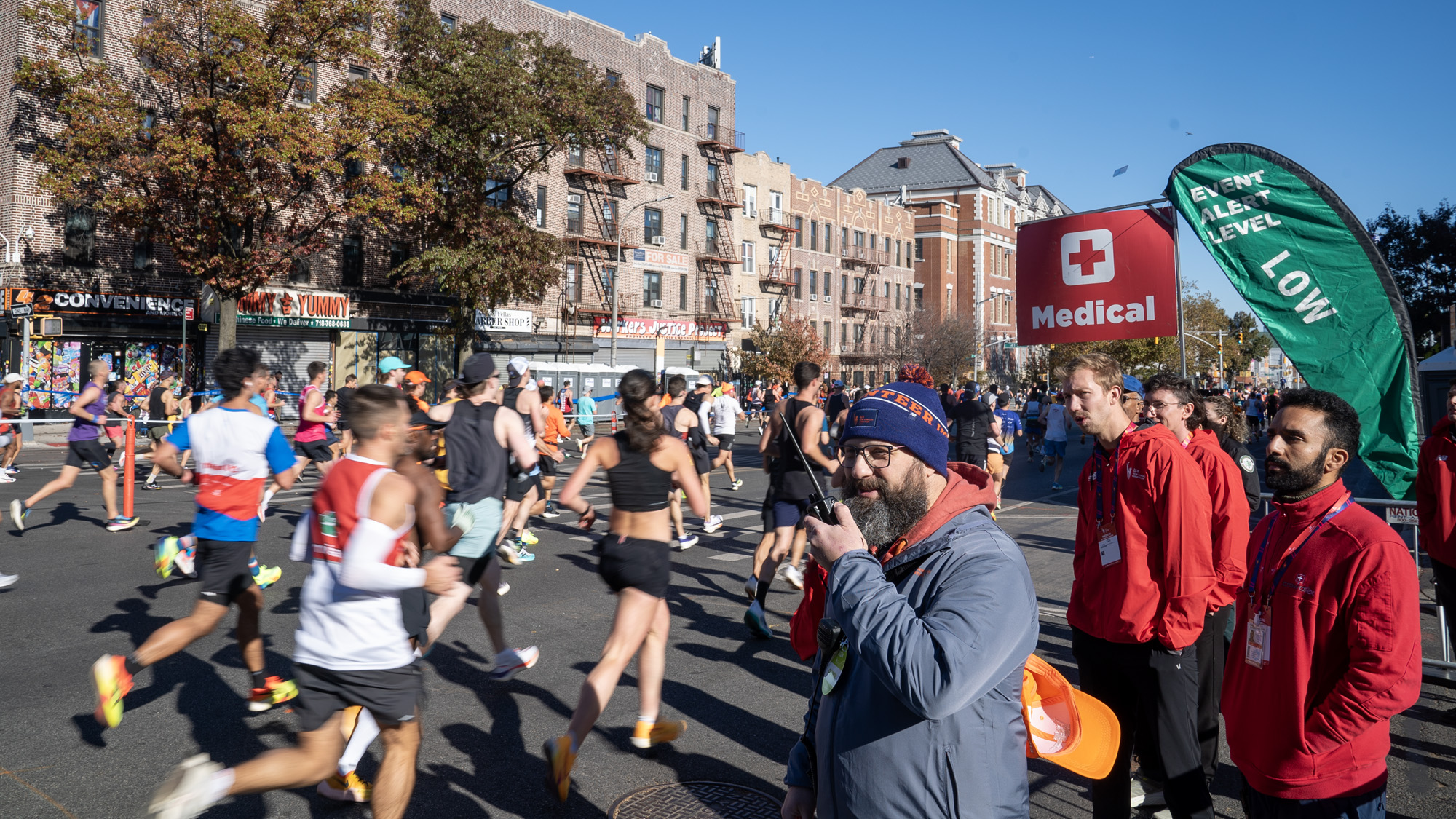 A bearded man in a hat holds a walkie-talkie as runners pass