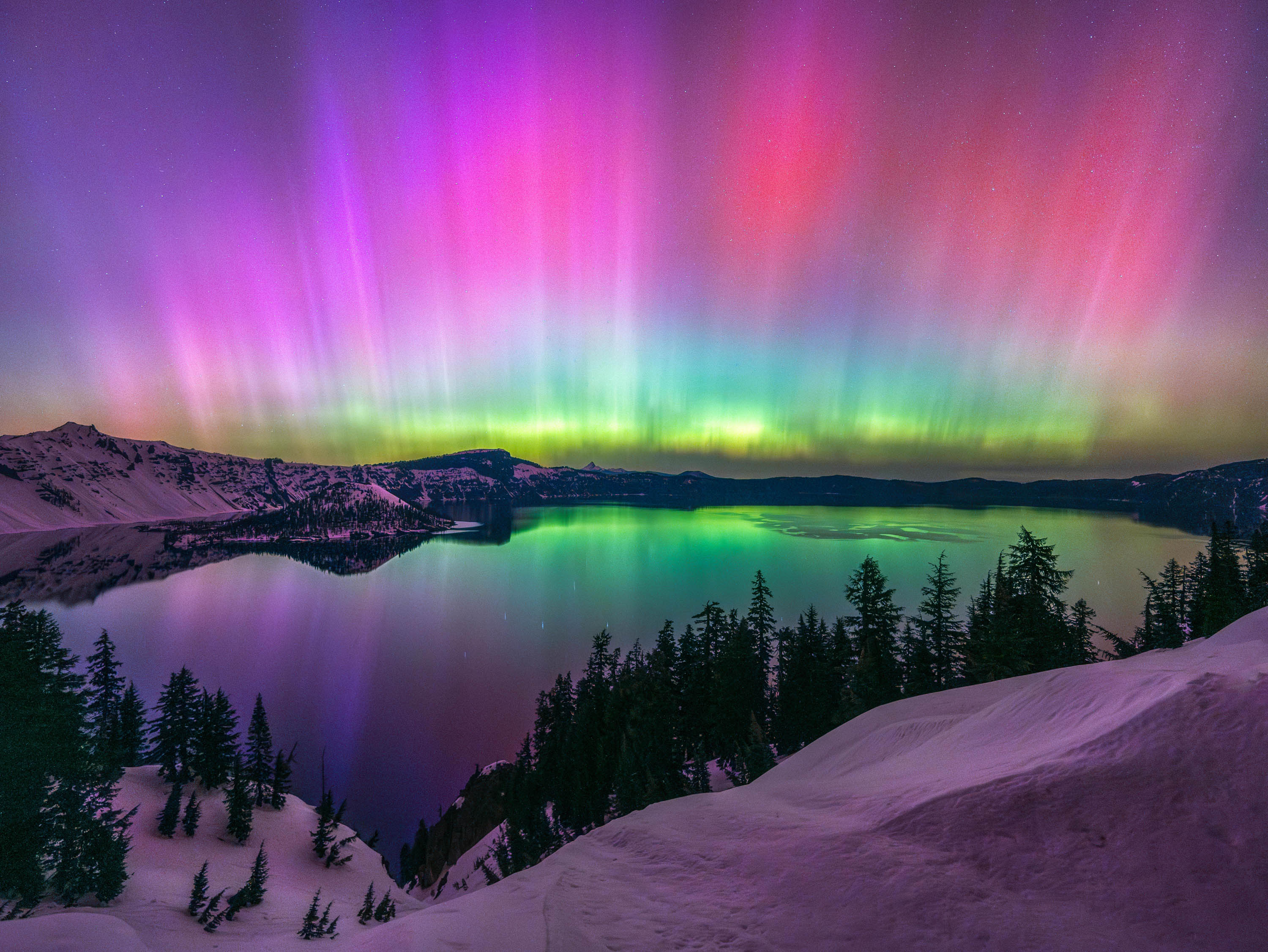 a rainbow aurora appears to shoot out from the horizon in front of a lake and hills covered in snow