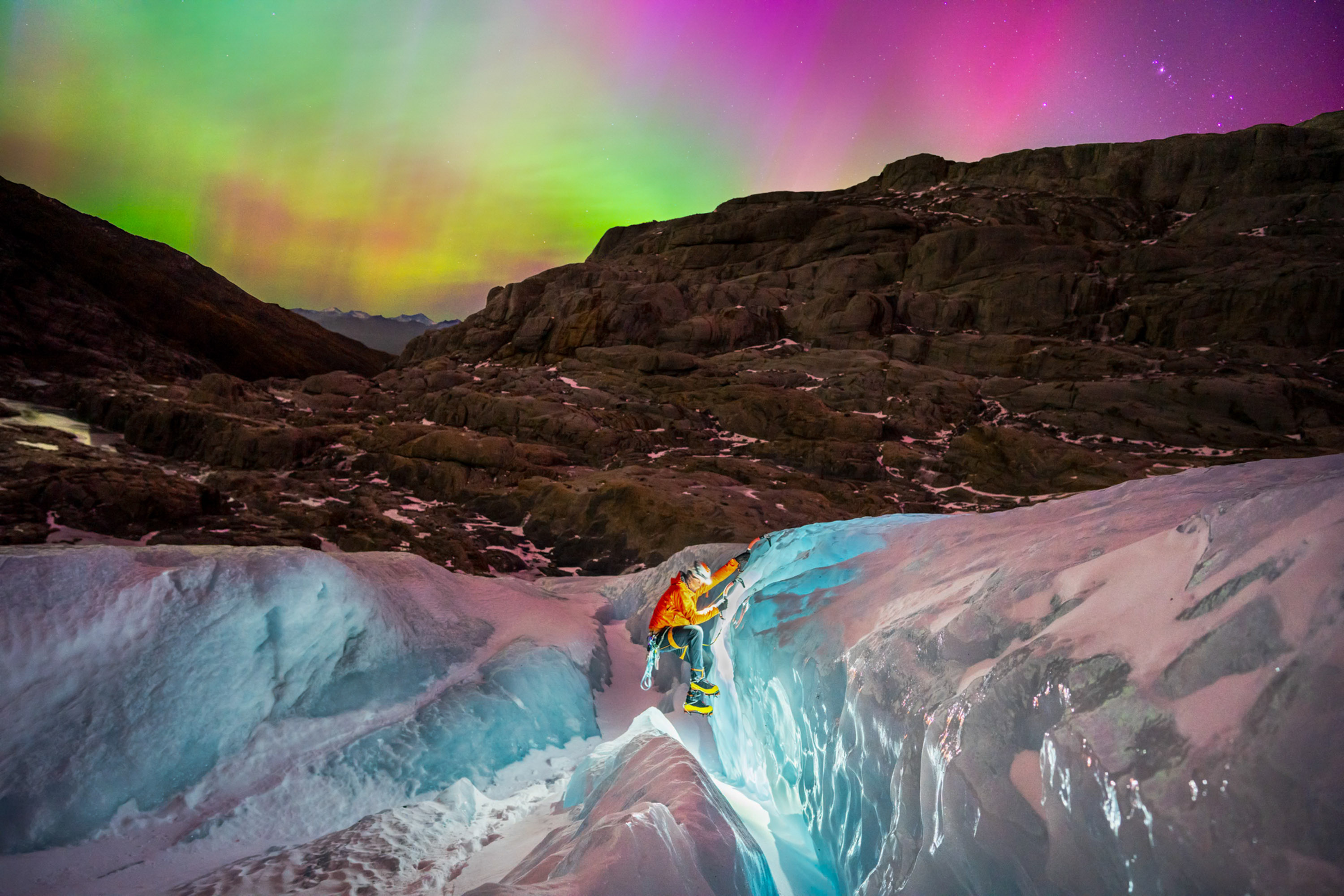 a glacier hiker climbs in the foreground with a headlamp as a rainbow colored aurora dances above