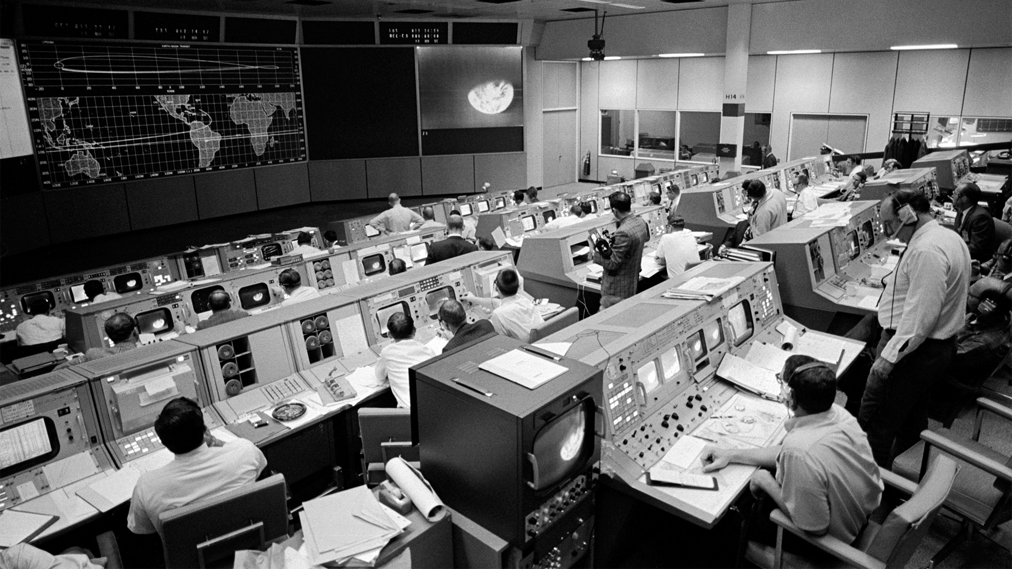 a black and white photograph of NASA mission control. several men are sitting at various screens and work stations