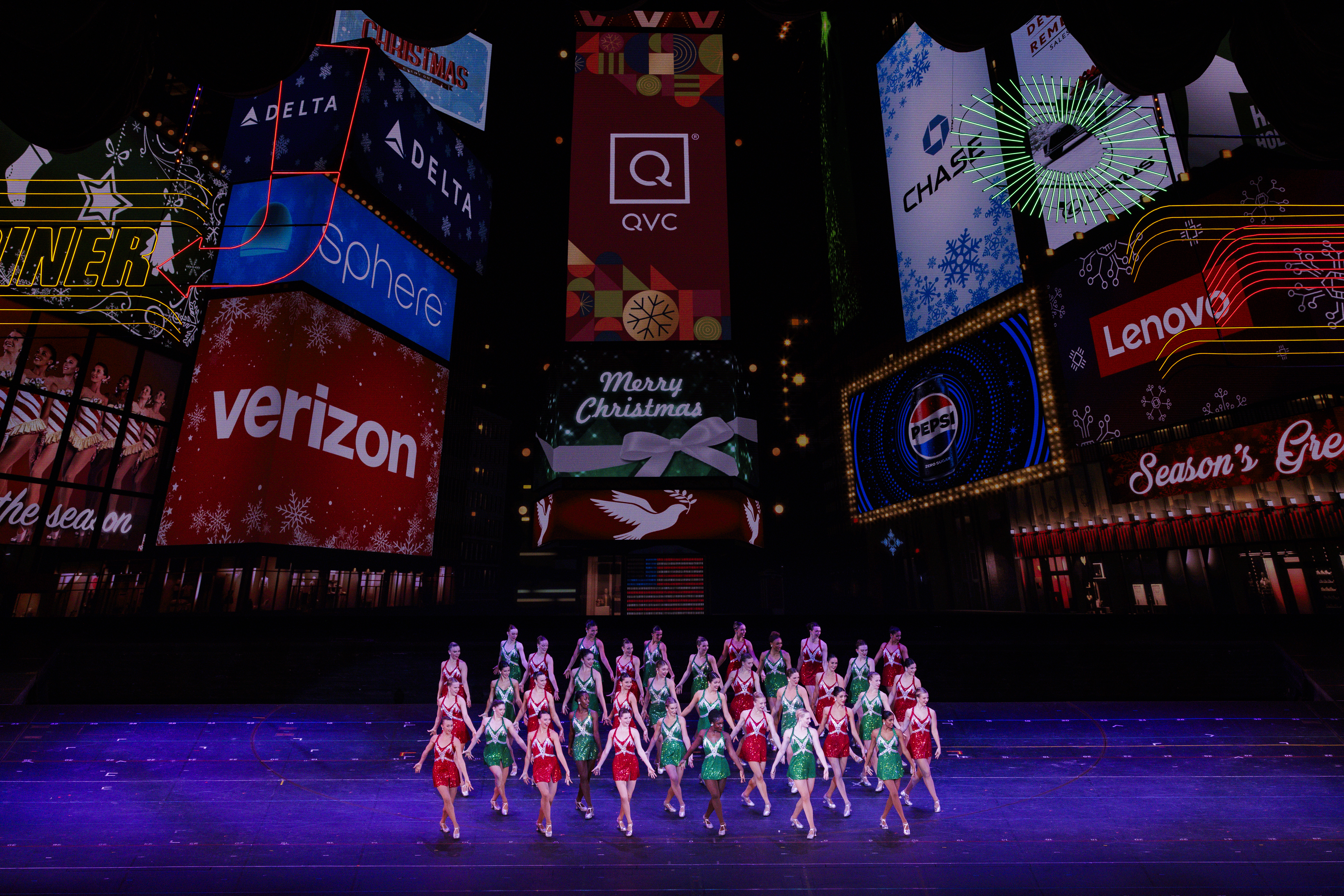 the rockettes on stage in red and green dresses and times square billboards projected behind them 