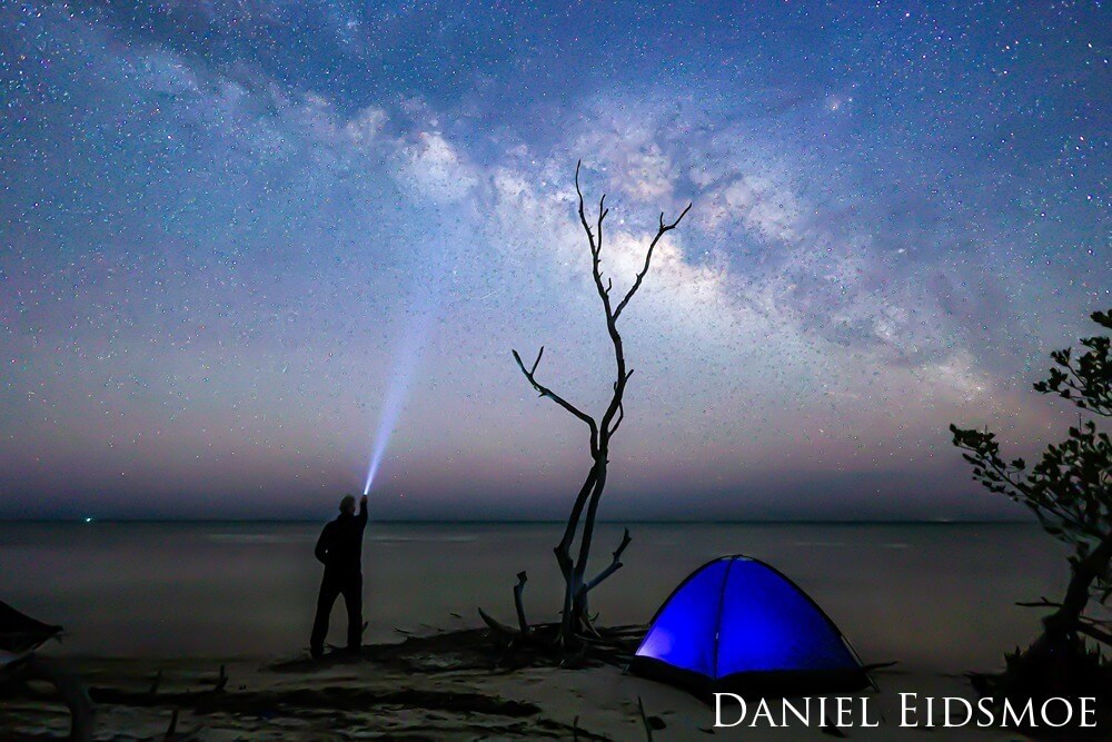 a camper with a flashlight shining up. the milky way is seen in the sky