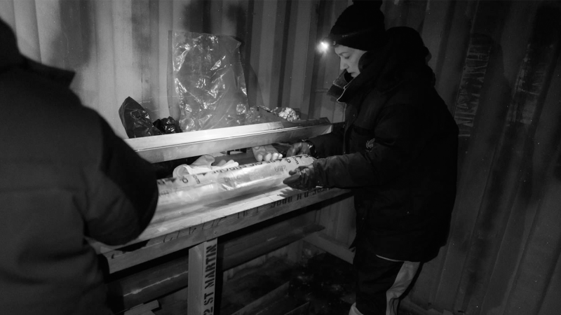 a scientist weather a larger winder coat and hat examines a cylindrical piece of ice