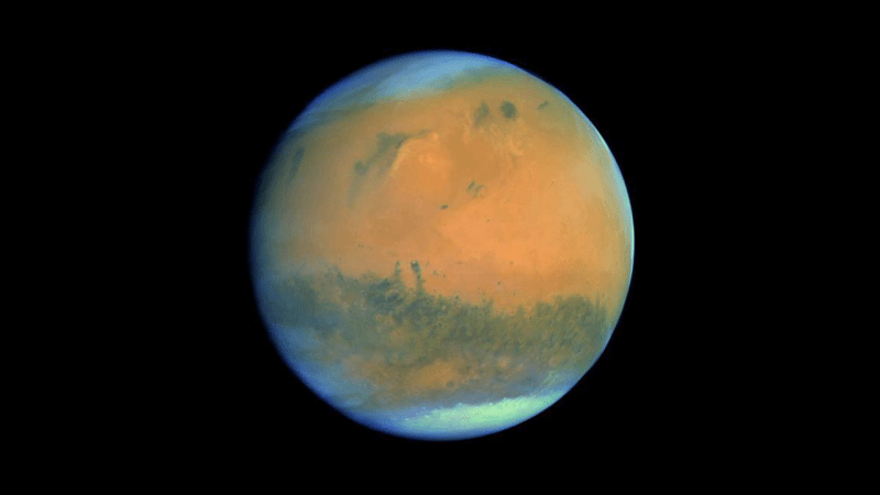 The full disc of Mars is seen with the polar ice caps slightly off centre to the top left and bottom right. Clouds wrap around the planet’s curved horizons. Dark surface markings are clearly seen against the characteristic red tones of the dusty martian surface.