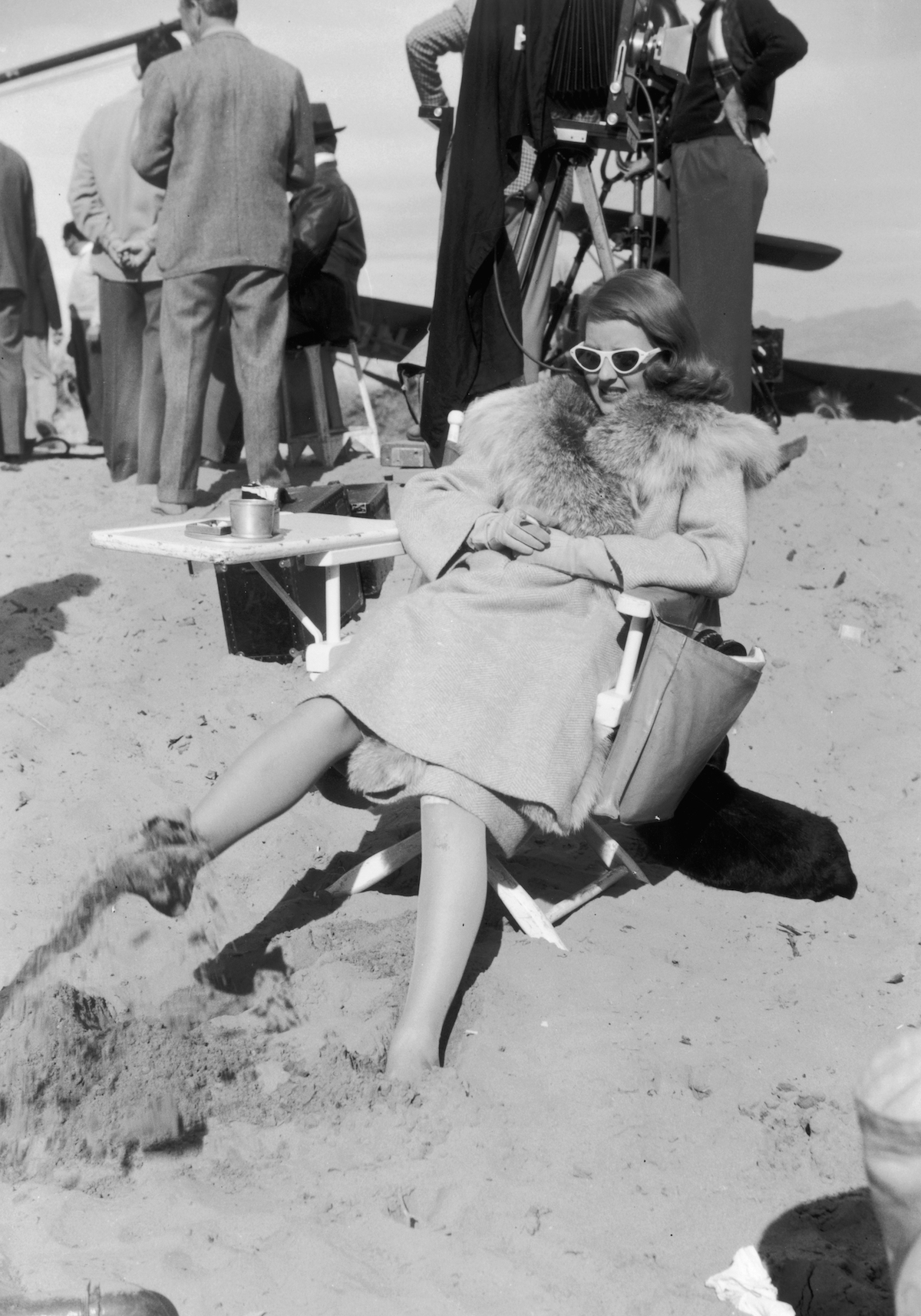  Bette Davis (1908-1989), sitting in a director's chair in the desert, holds a cigarette and plays in the sand with her foot during a break on the set of director William Keighley's film, 'The Bride Came C.O.D.,' Death Valley, California. Davis is wearing a fur-trimed coat, gloves, white-framed cat eye sunglasses, and stockings. 