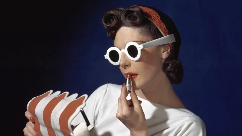 Model Muriel Maxwell in white sunglasses putting on lipstick, wearing red-white-and-blue turban, andv holding a red-and-white striped bag *** Local Caption ***